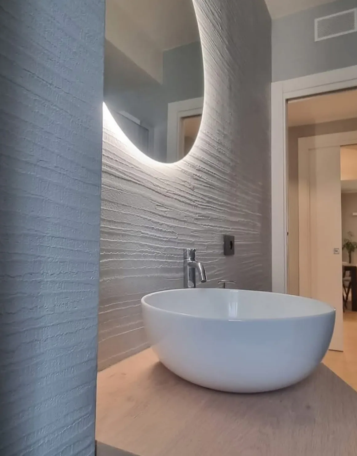 Textured plaster bathroom wall with a backlit round mirror and freestanding bath